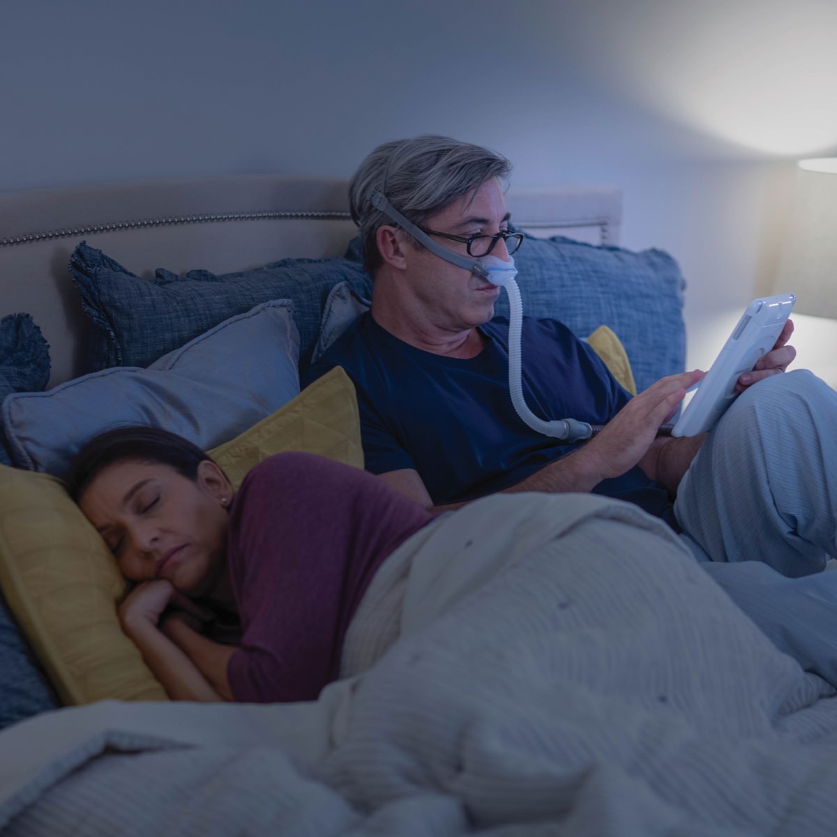 Man Sitting in Bed Wearing His Nasal CPAP Mask Next to His Partner