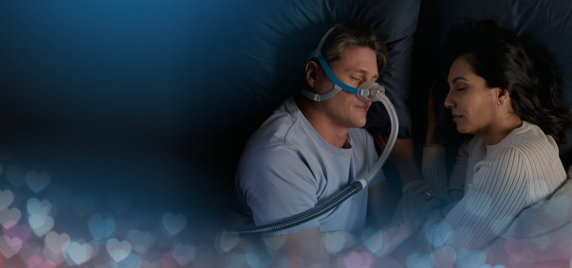 Person using a CPAP machine with a partner in a dimly lit room, surrounded by heart-shaped lights.