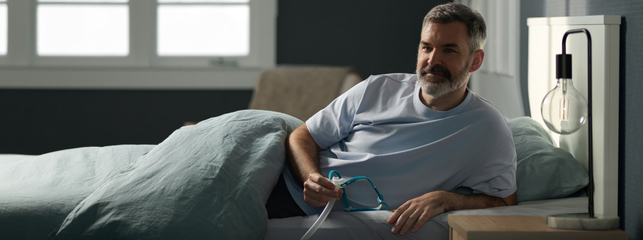 Man Lying Propped Up On His Side Holding His Fisher & Paykel Nova Micro Nasal Pillow CPAP Mask W/ Headgear