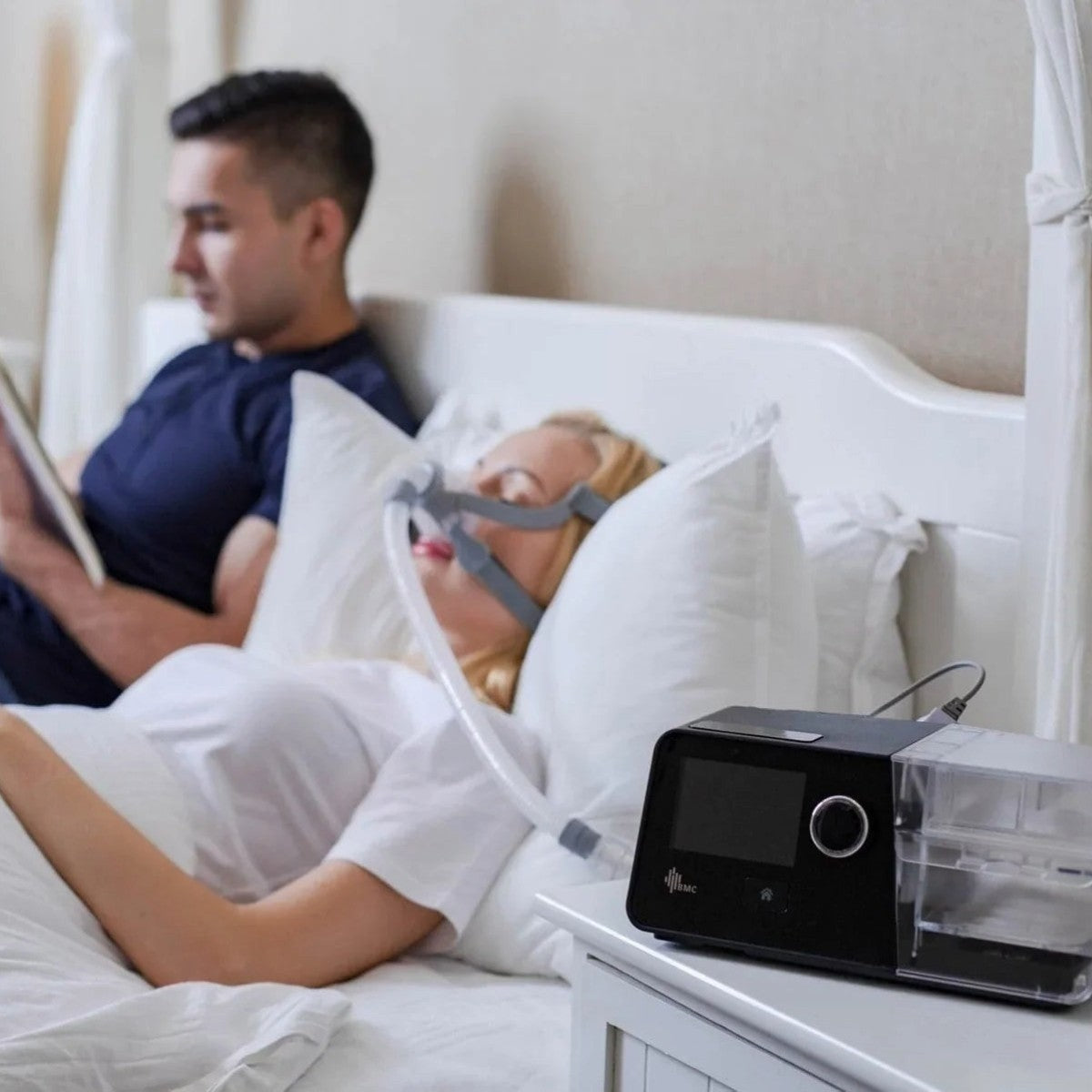 Person using a CPAP machine with another person in bed, likely for sleep apnea treatment.