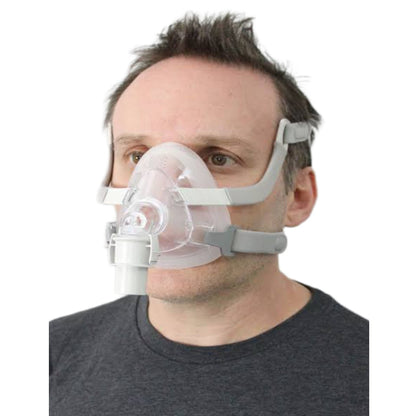 A man wearing a clear CPAP mask with gray straps and a white connector, shown against a plain white background.